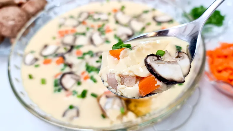 lets eat A spoon holding up a bite of Smooth Steamed Egg with bits of chicken, mushrooms and carrots.